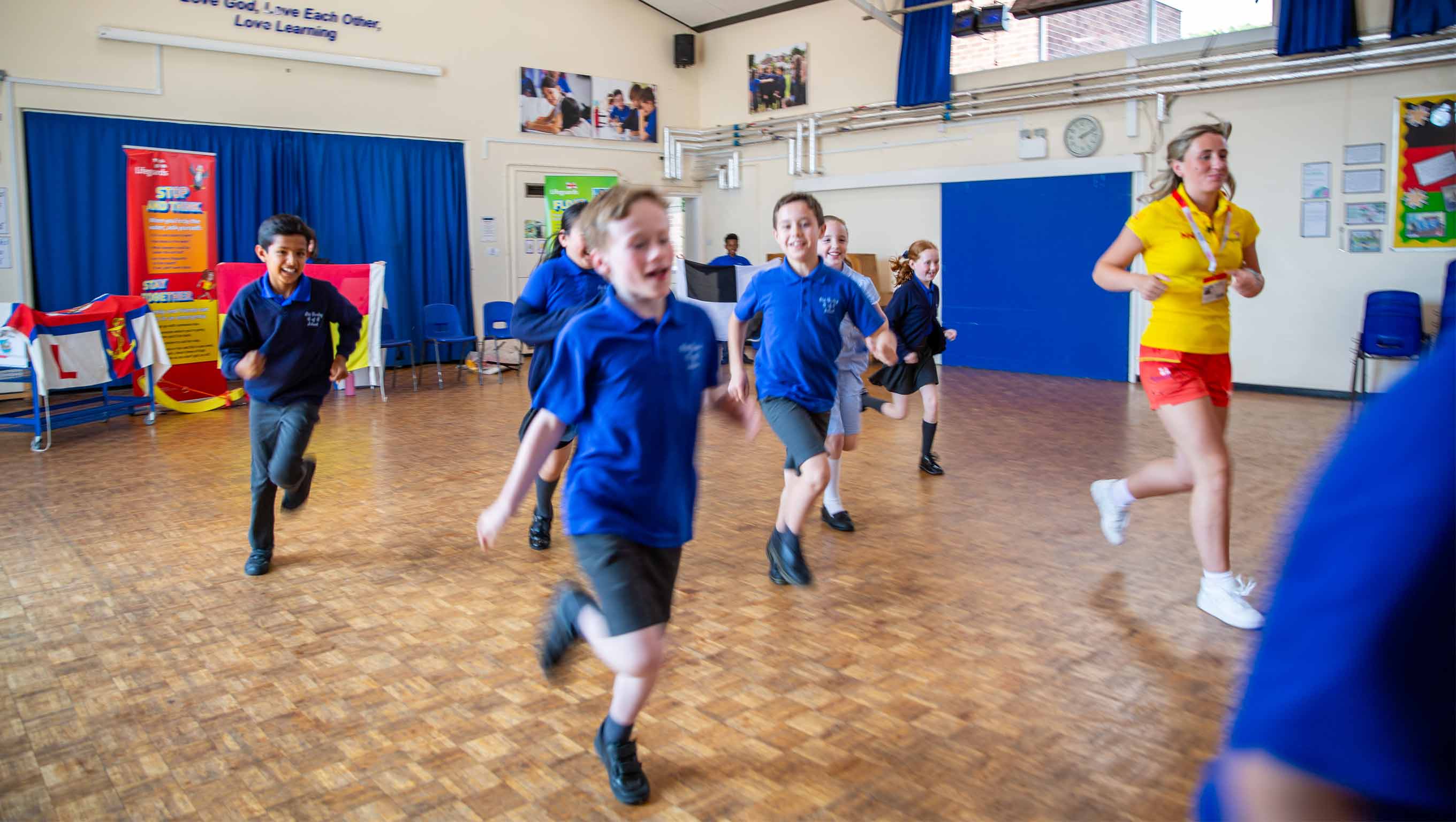 A group of school children run across an assembly hall following a female lifeguard wearing yellow and red lifeguard kit