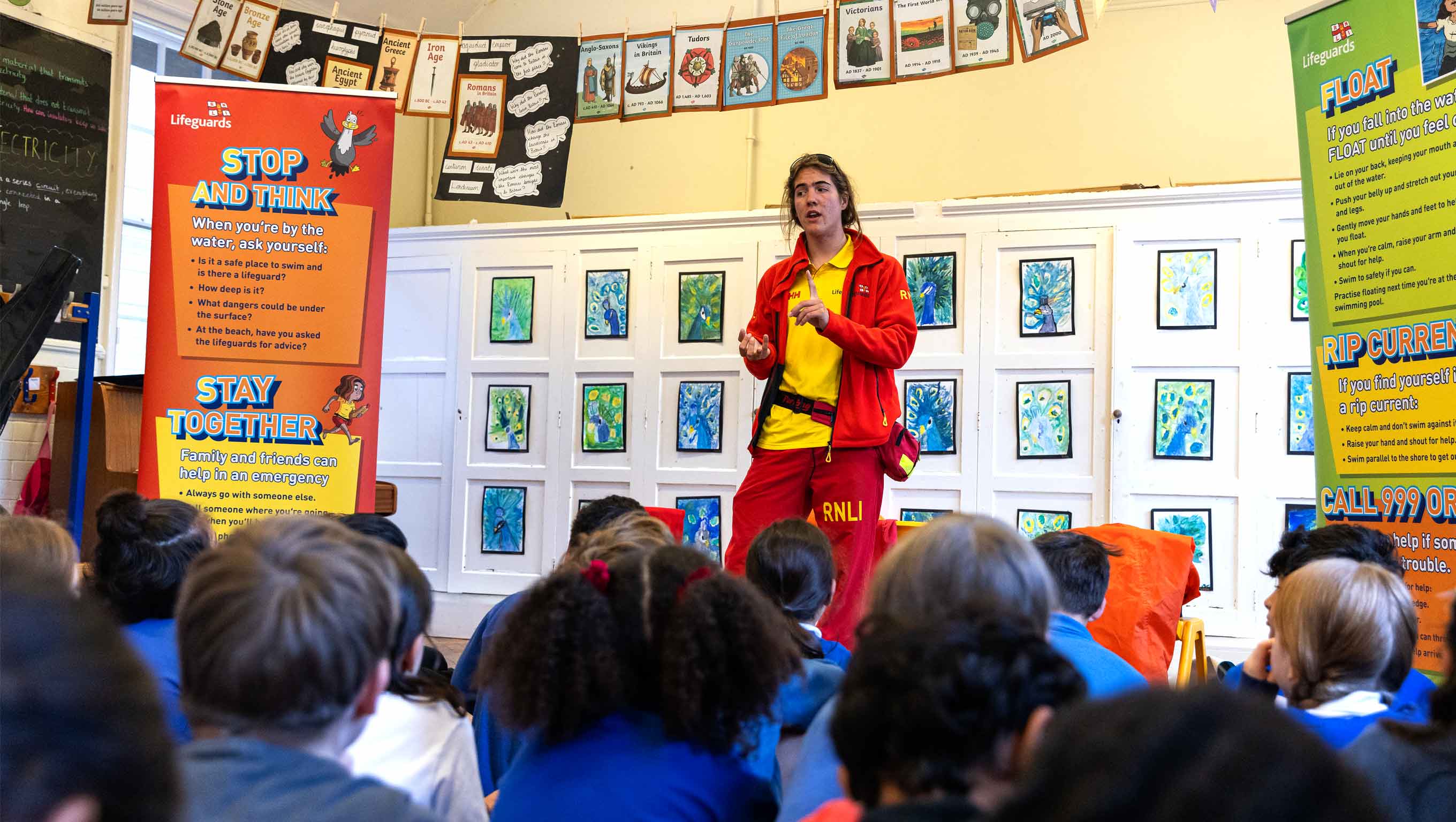 A lifeguard stands in front of a crowd of school children giving a presentation on water safety. He is wearing a red lifeguard fleece, yellow polo, and red shorts. On either side of him are sea safety pull-up banners.