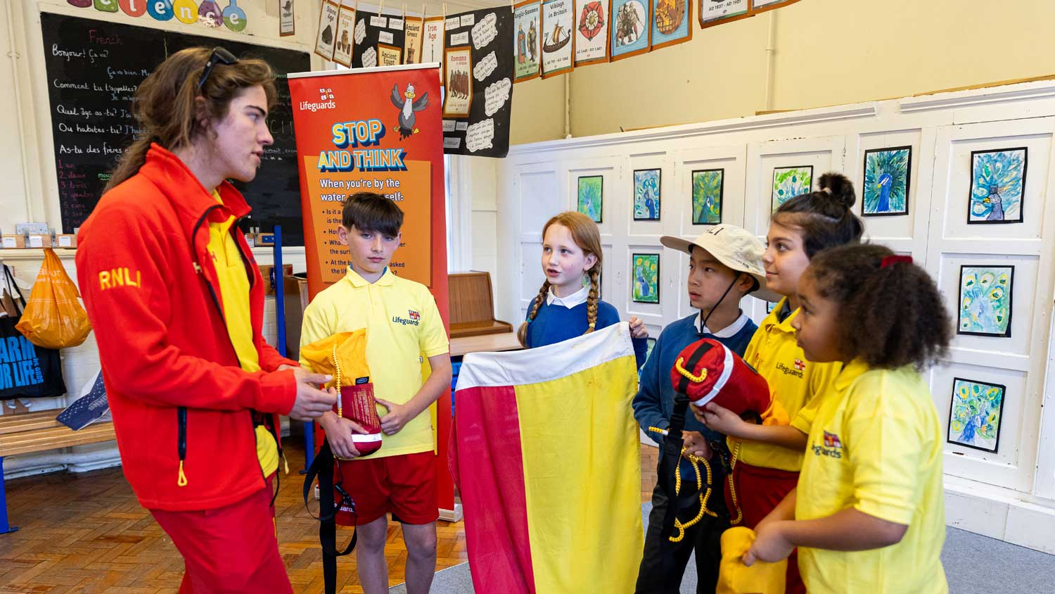 A female RNLI lifeguard wearing red lifeguard jacket, yellow polo shirt and red shorts stands in an assembly hall chatting with a group of five students from a mix of different backgrounds