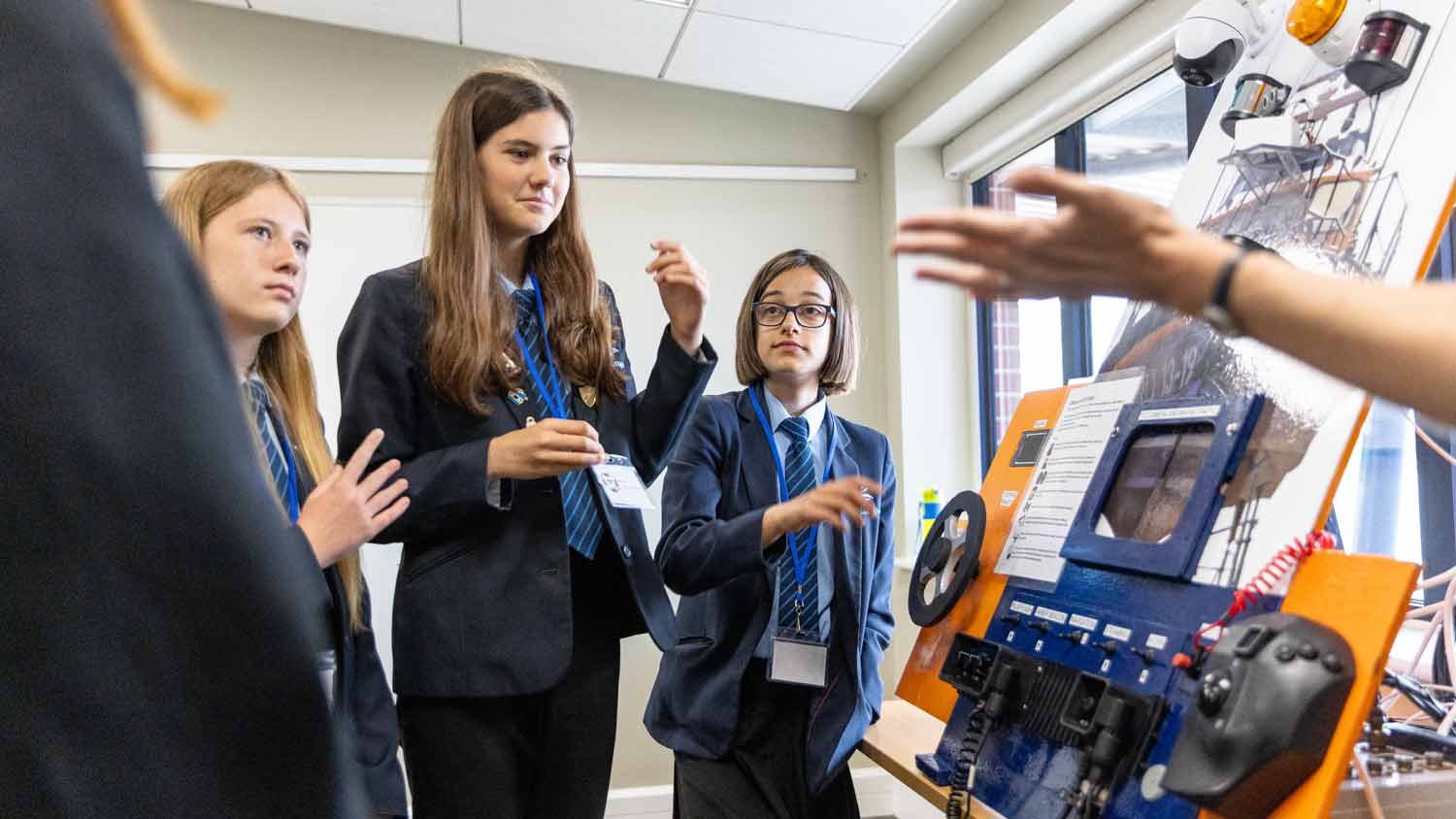 Three girls in school uniform stand next to a display of RNLI lifeboat engineering with various knobs gears and dials as an instructor explains how it works