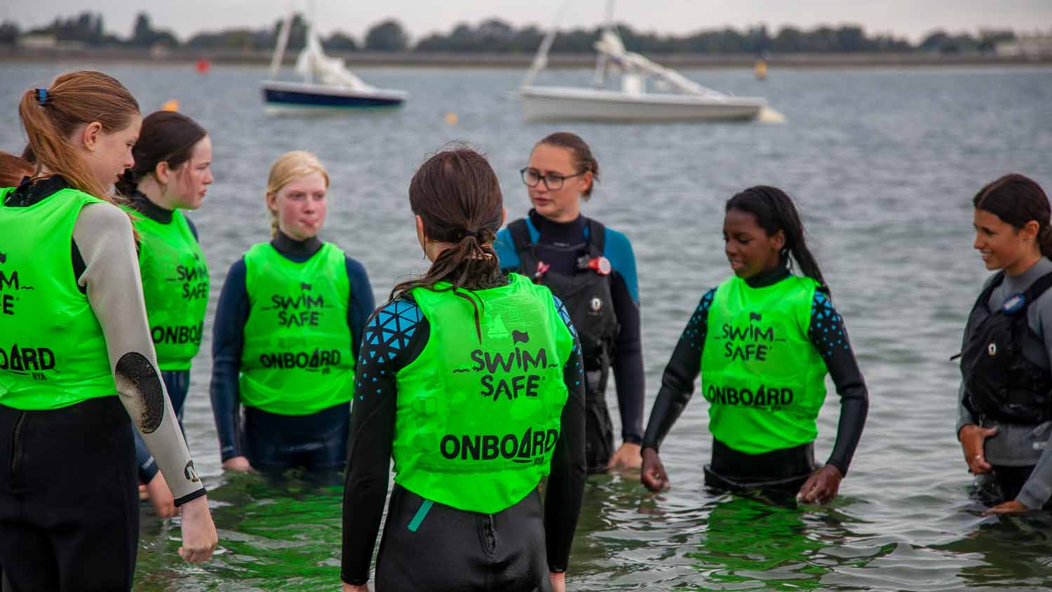 A group of girls in bright green Swim Safe buoyancy aids stand in the sea during a Swim Safe session. Two instructors in black buoyancy aids stand nearby.