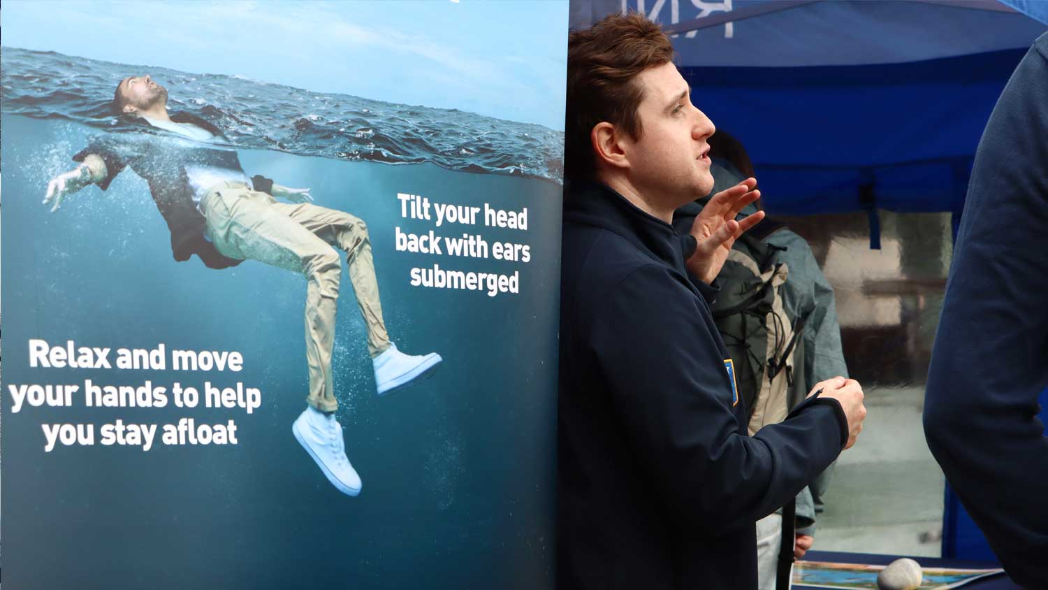 An RNLI water safety volunteer talks through some safety tips as he stands next to a pull-up board with Float to Live advice written on it