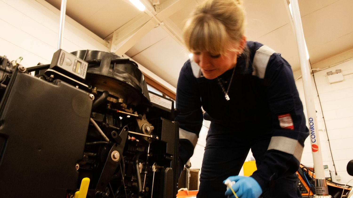 A woman in dark blue overalls and light blue gloves is intently focused on working on a large black engine