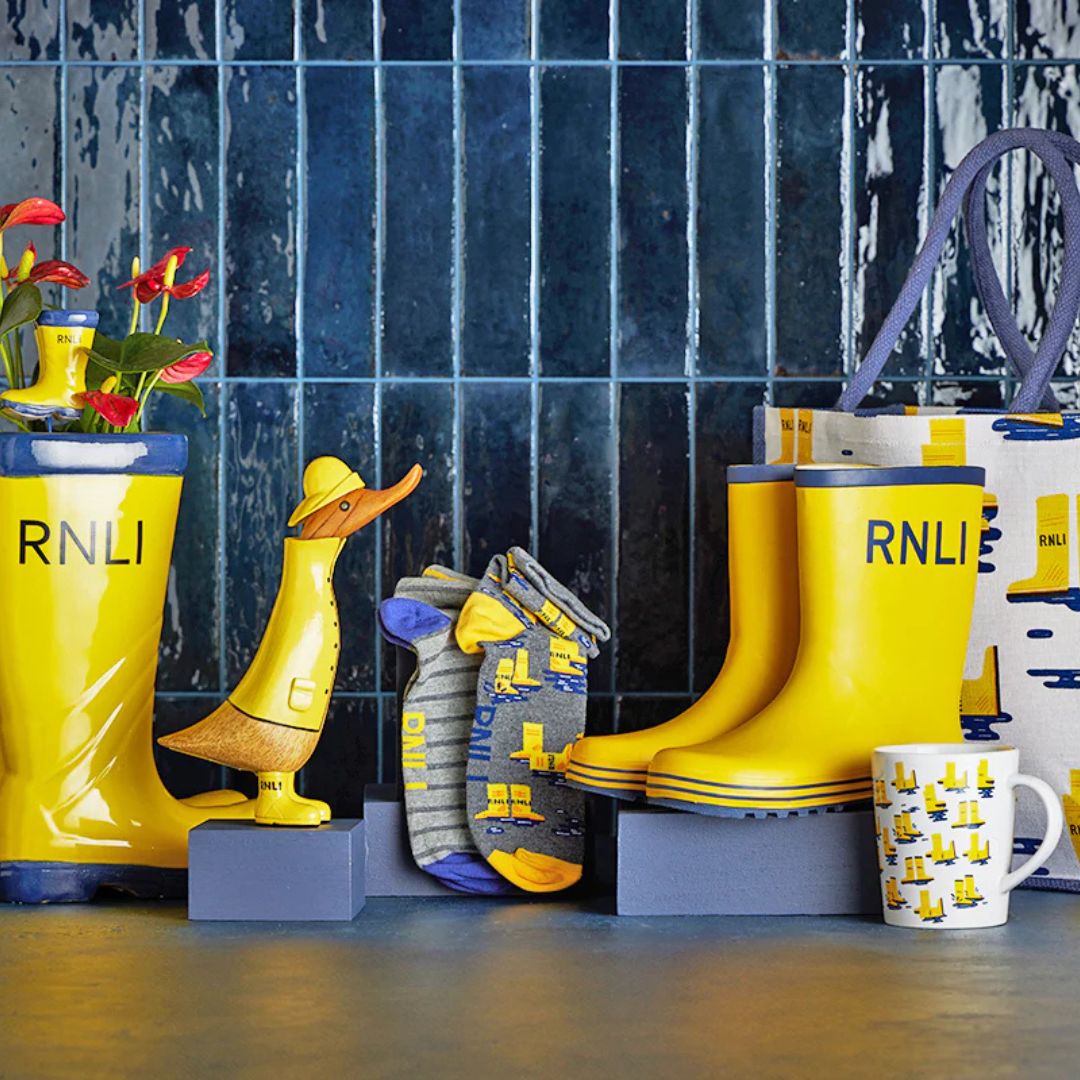 A collection of Mayday items including a wooden duck wearing a yellow raincoat, hat, and wellies, a pair of socks, a mug, a shopping bag. 