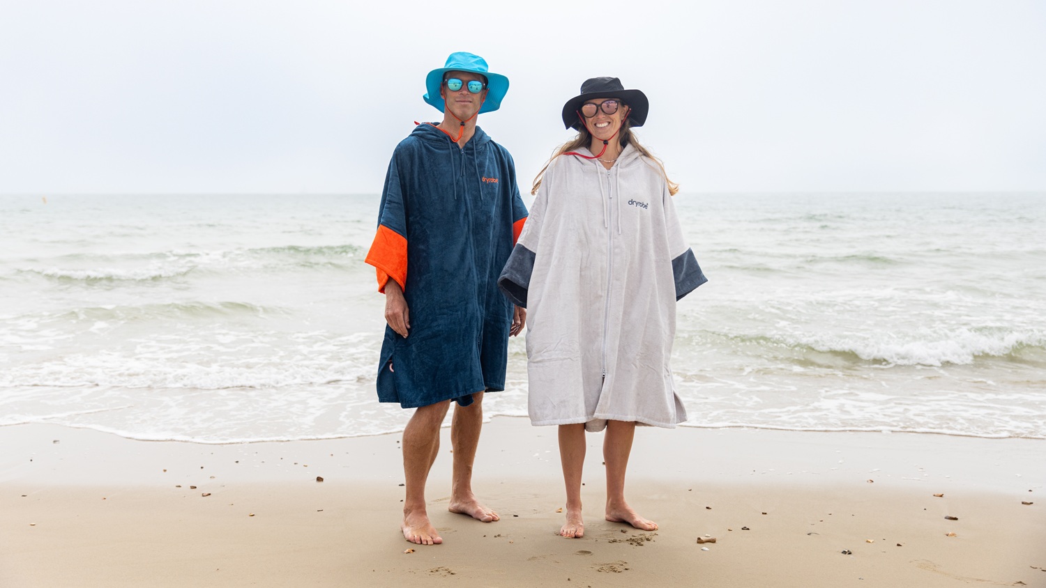 A man and woman standing together, barefoot, on a wet sandy shoreline wearing towelling dryrobes, dryrobe brimmed hats and sunglasses.