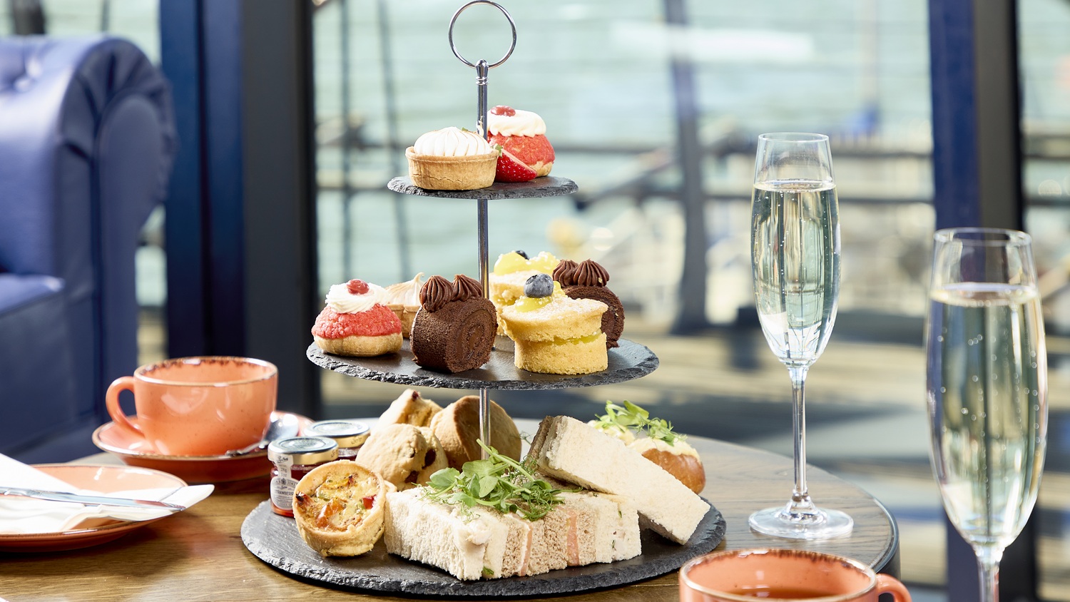 A selection of sandwiches, cakes and scones on a three-tiered stand with two cups of tea on saucers and two flutes of Champagne.