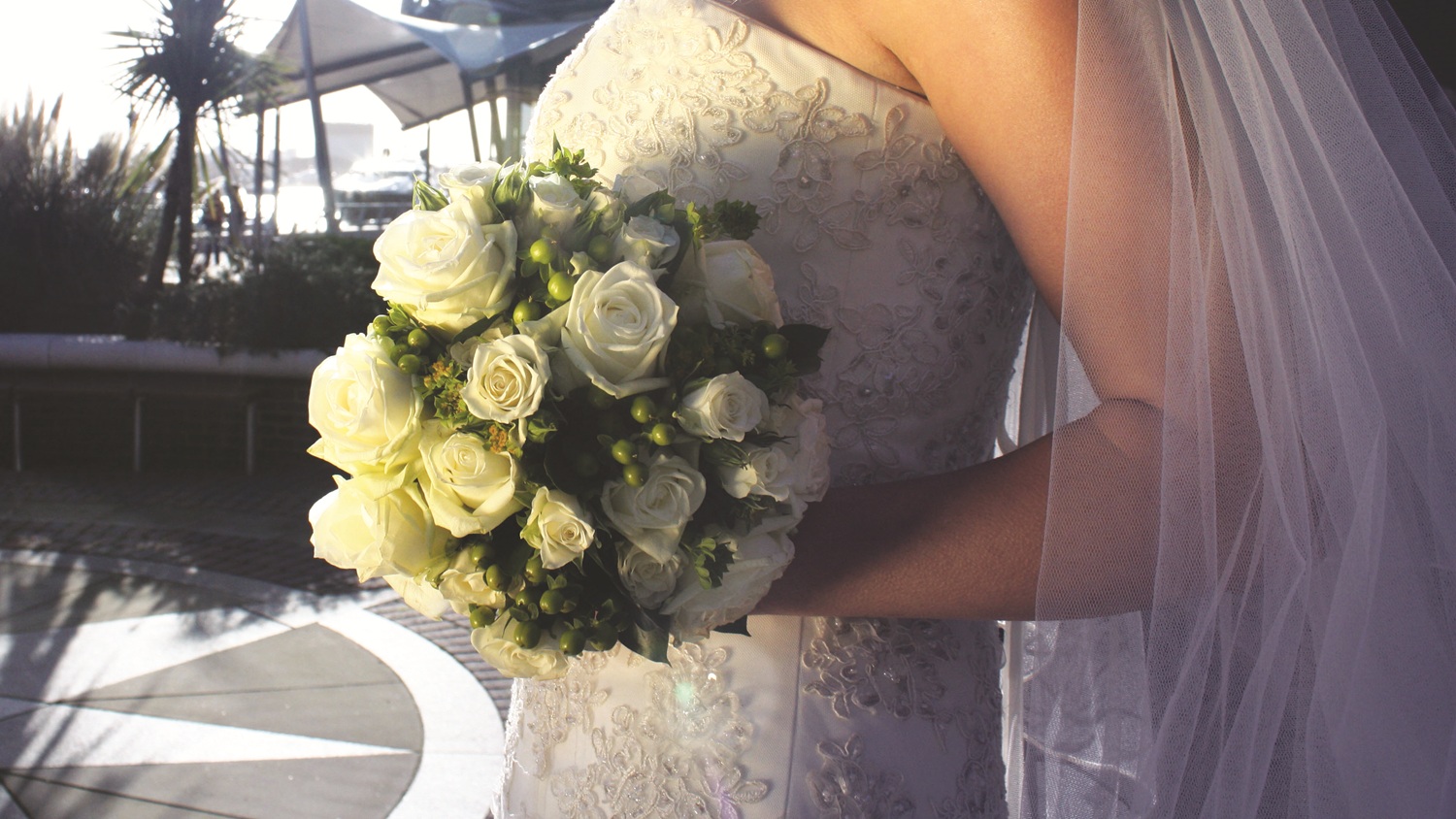 A zoomed-in, side-on view of a bride’s bouquet as she holds it by her waist on the sea terrace at the RNLI College. The bride is wearing an embellished white dress with a veil and her bouquet is made up of white roses. Her face is not visible.