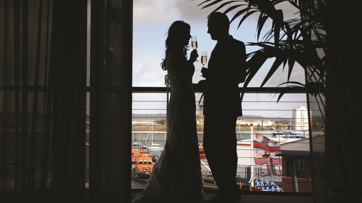 The silhouette of a wedding couple as they stand facing each other on the balcony of the Harbour View Suite at the RNLI College. They are each holding a glass of Champagne.