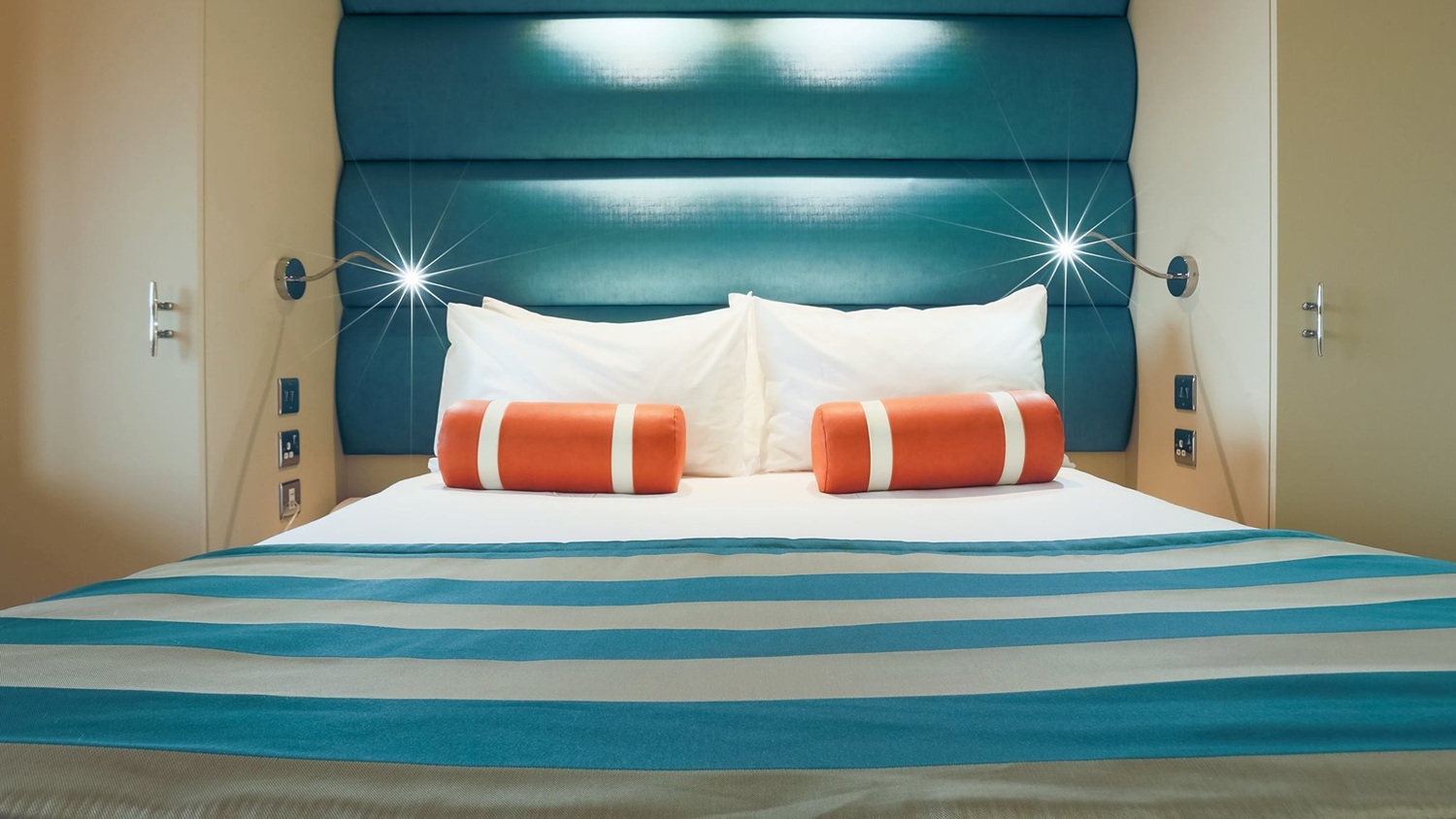 A modern bedroom at the RNLI College with a nautical striped double bed, barrel-shaped cushions and bedside wall lamps.