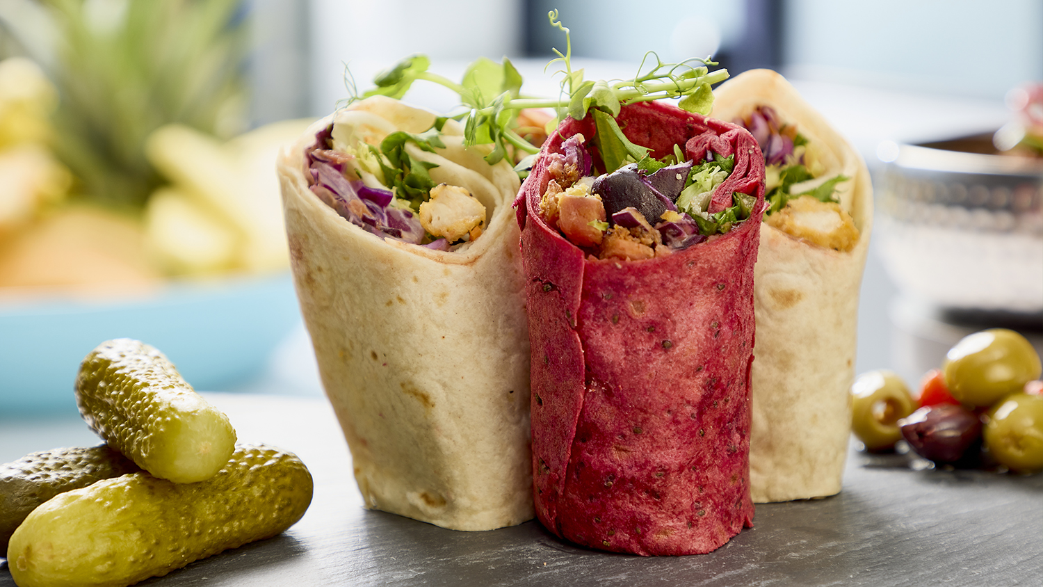 A selection of filled wraps alongside gherkins and olives on a black slate serving dish.